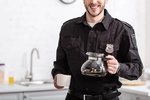 Vista parcial de policía sonriente sosteniendo jarra de vidrio de café filtrado en la cocina - foto de stock
