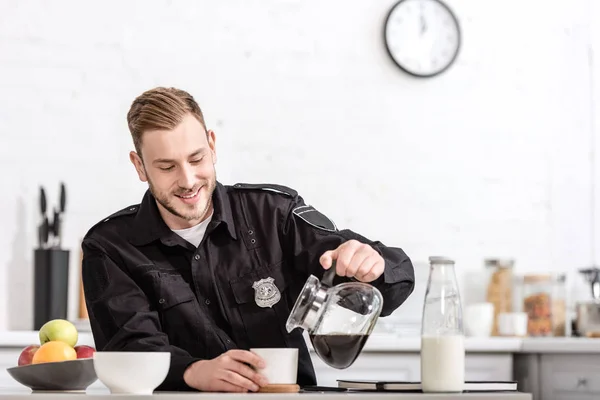 Oficial de policía sonriente vertiendo café filtrado de maceta de vidrio en la cocina - foto de stock