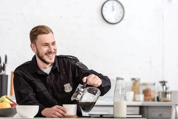 Oficial de policía feliz verter café filtrado de maceta de vidrio en la cocina - foto de stock