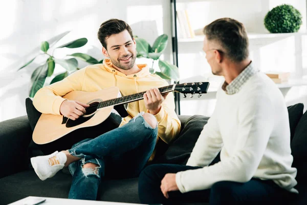 Smiling son playing acoustic guitar for mature father on weekend at home — Stock Photo