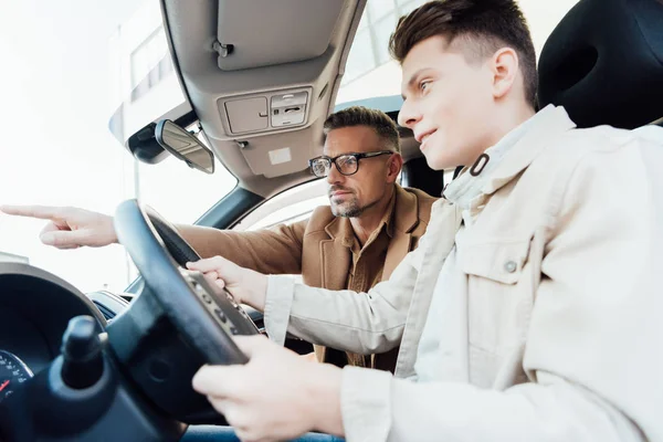 Seitenansicht eines hübschen Vaters, der auf etwas zeigt, während er Sohn Autofahren beibringt — Stockfoto