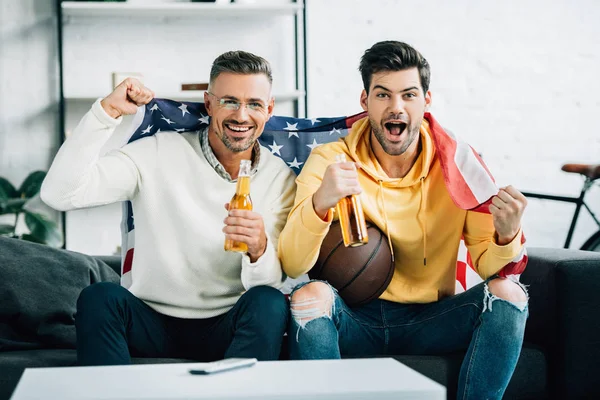 Excited son and mature father wrapped in united states flag watching basketball game on weekend at home — Stock Photo