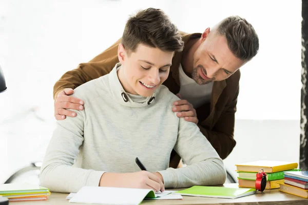 Padre sorridente abbracciare figlio adolescente mentre fa i compiti e scrivere qualcosa — Foto stock