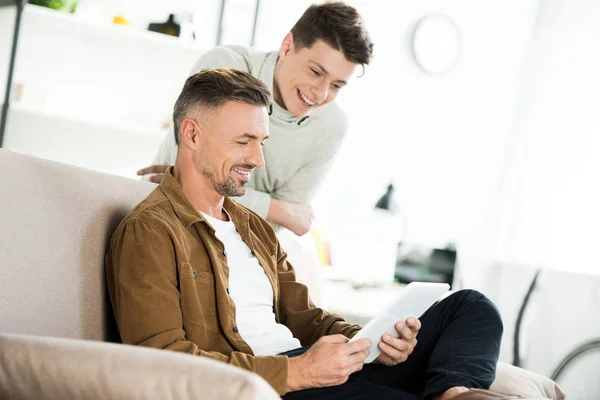 Sonriente padre e hijo adolescente mirando tableta en la sala de estar - foto de stock
