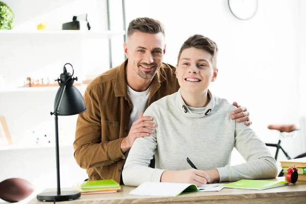 Lächelnder Vater umarmt Teenager-Sohn, während er Hausaufgaben macht und in die Kamera schaut — Stockfoto