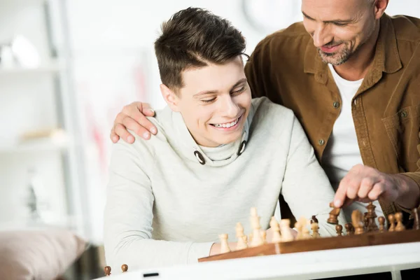 Sonriente padre abrazando feliz adolescente hijo y jugando ajedrez en casa - foto de stock