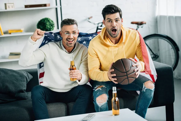 Surprised son and mature father wrapped in united states flag watching basketball game on weekend at home — Stock Photo