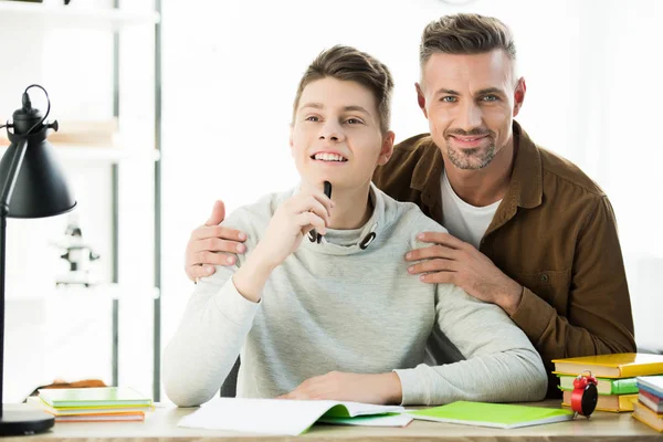 Sonriente padre abrazando pensativo adolescente hijo mientras él haciendo tarea - foto de stock