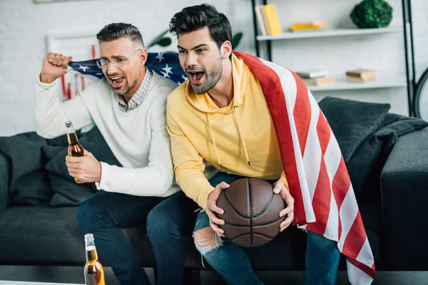 Збуджений син і зрілий батько, загорнутий в єдиний прапор штатів, дивиться баскетбольну гру і кричить у вихідні вдома — стокове фото