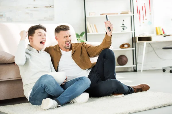 Emocionado padre e hijo adolescente viendo partido en la televisión y sosteniendo tazón de palomitas de maíz en casa - foto de stock