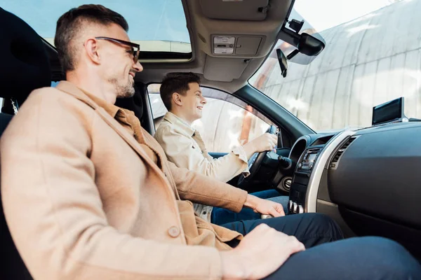 Side view of smiling handsome father teaching teen son driving car — Stock Photo