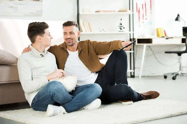 Cheerful father and teen son watching tv and holding bowl of popcorn at home — Stock Photo