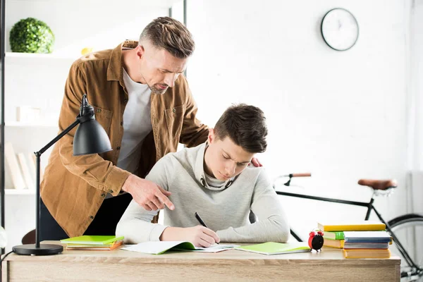 Father pointing on something to teen son while he writing in copybook — Stock Photo