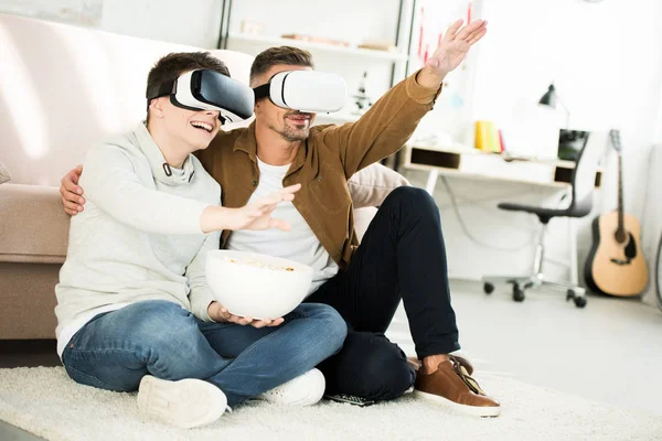 Padre e hijo adolescente viendo algo con auriculares de realidad virtual en casa - foto de stock