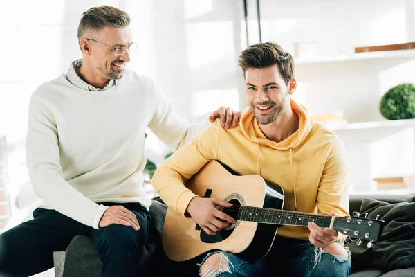 Alegre hijo tocando la guitarra acústica para padre maduro en fin de semana en casa - foto de stock