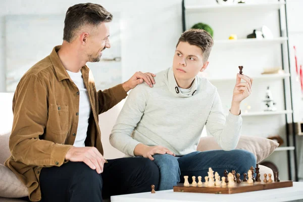 Lächelnder Vater umarmt Teenager-Sohn, während er zu Hause Schachfigur hält — Stockfoto