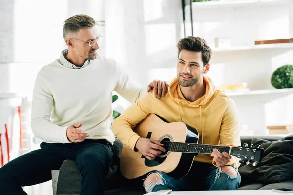 Smiling son playing guitar for mature father on weekend at home — Stock Photo