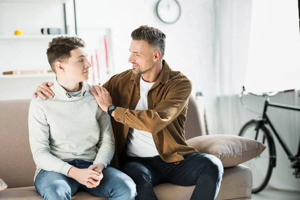 Padre abrazando adolescente hijo en sofá en casa - foto de stock