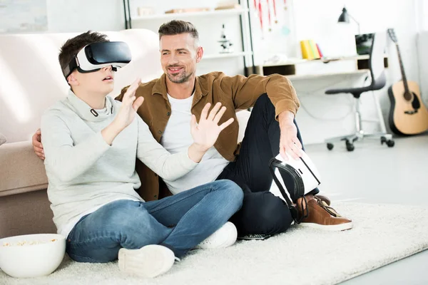 Pai alegre olhando para o filho adolescente assistindo algo com fone de ouvido realidade virtual em casa — Fotografia de Stock