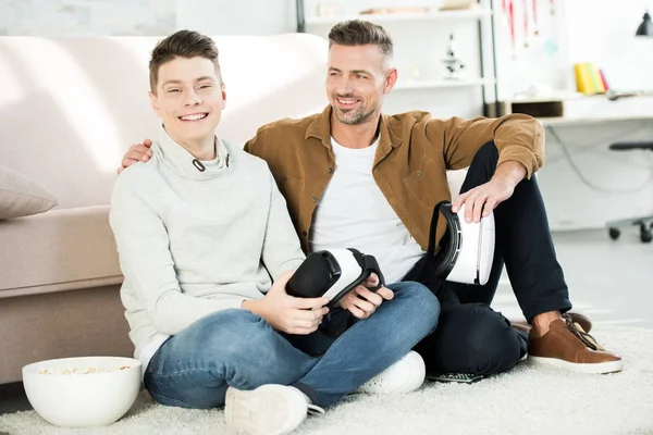 Happy father and teen son holding virtual reality headsets at home — Stock Photo