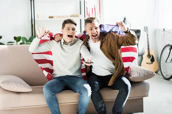 Excited father and teen son wrapped in united states flag screaming on sofa during watching sport match — Stock Photo