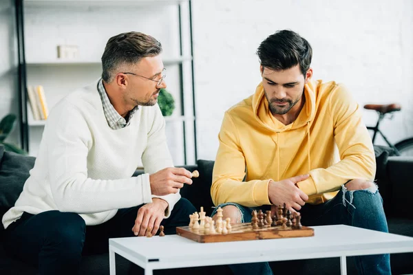 Hijo y padre maduro jugando ajedrez juntos en fin de semana en la sala de estar - foto de stock