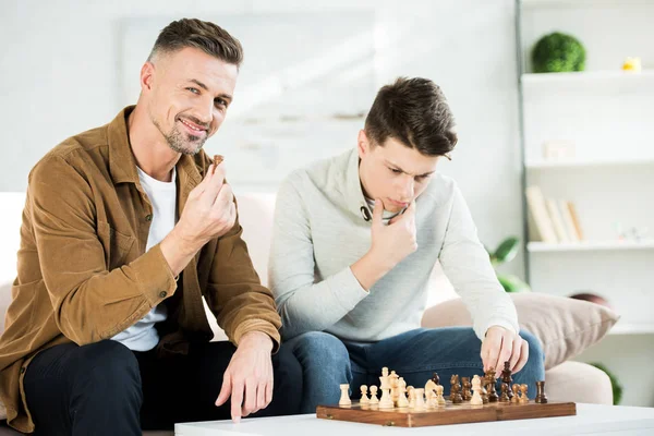 Padre pensativo e hijo adolescente jugando ajedrez en la sala de estar - foto de stock