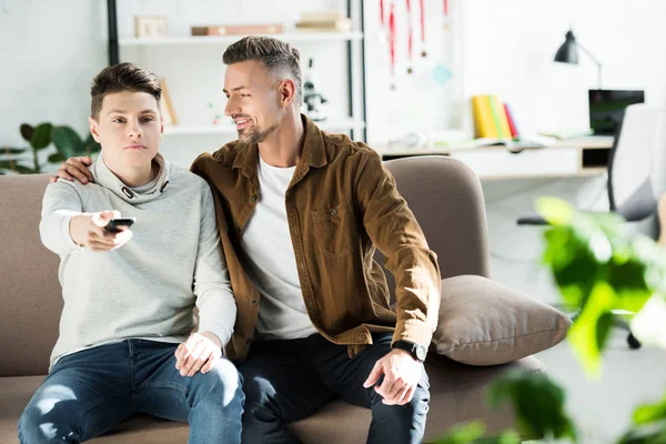 Sonriente padre abrazando hijo adolescente, chico sosteniendo control remoto en casa - foto de stock