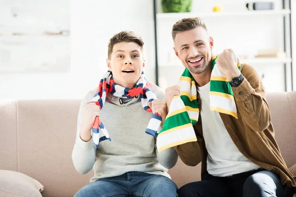 Padre emocionado e hijo adolescente en bufandas viendo deporte partido y gesto en casa - foto de stock