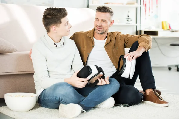 Sorrindo pai e filho adolescente segurando fones de ouvido realidade virtual em casa e olhando um para o outro — Fotografia de Stock
