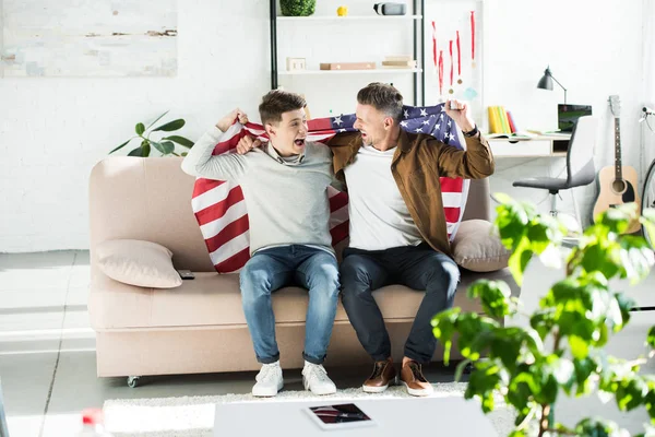 Heureux père et adolescent fils enveloppé dans drapeau des États-Unis assis sur canapé et criant pendant le match de sport — Photo de stock