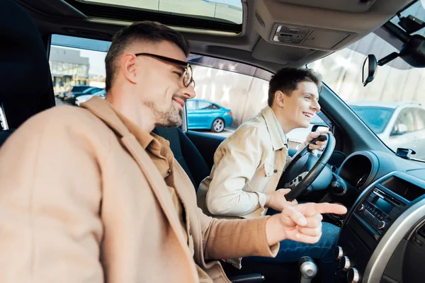 Lächelnder schöner Vater zeigt auf etwas auf der Straße, während er Teenager-Sohn Autofahren beibringt — Stockfoto