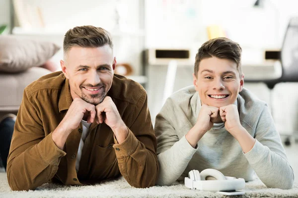 Padre sorridente e figlio adolescente sdraiato sul tappeto e guardando la fotocamera a casa — Foto stock