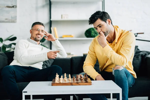 Filho pensativo e sorrindo pai maduro jogando xadrez juntos no fim de semana em casa — Fotografia de Stock