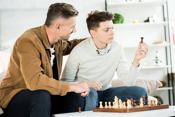 Padre abrazando hijo adolescente mientras mira la figura de ajedrez en casa - foto de stock