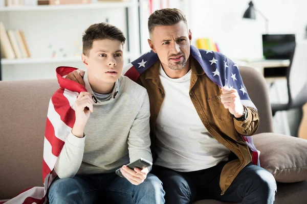 Серьезный отец и сын-подросток, завернутые в флаг США, сидят на диване и смотрят спорт дома — стоковое фото