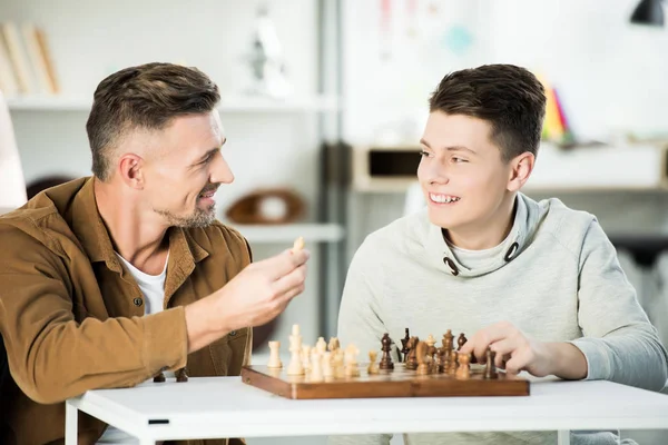 Sonriente padre e hijo adolescente jugando ajedrez en casa y mirándose el uno al otro — Stock Photo