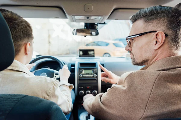 Back view of handsome father pointing on something while teaching teen son driving car — Stock Photo