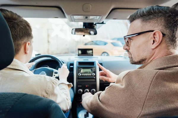 Vista posteriore di bel padre che punta su qualcosa mentre insegna adolescente figlio guida auto — Foto stock