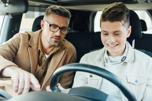 Schöner Vater zeigt auf etwas, während er lächelnden Teenie-Sohn Autofahren beibringt — Stockfoto