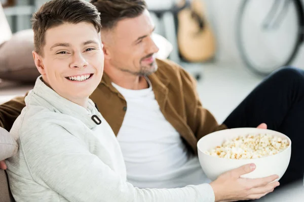 Ritratto di padre e felice figlio adolescente guardando la tv e tenendo ciotola di popcorn a casa — Foto stock