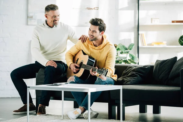 Feliz hijo tocando la guitarra acústica para padre maduro en fin de semana en casa - foto de stock