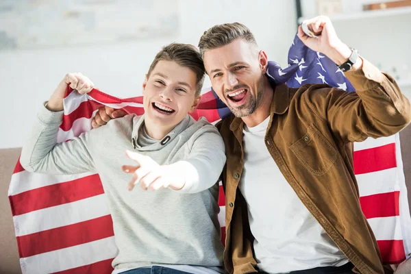 Excited father and teen son wrapped in united states flag screaming on sofa during sport match — Stock Photo