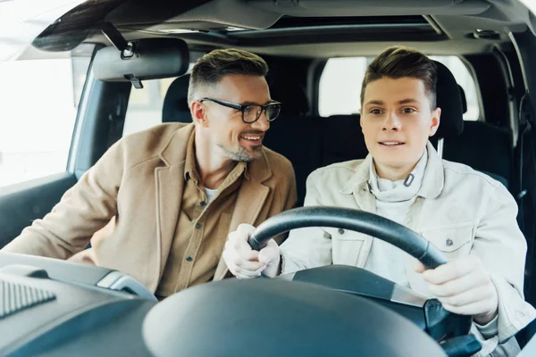 Lächelnd gut aussehender Vater lehrt Teenager-Sohn Autofahren und schaut ihn an — Stockfoto