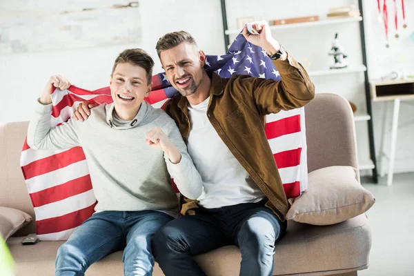 Feliz padre e hijo adolescente envuelto en la bandera de los estados unidos sentado en el sofá y viendo el partido deportivo - foto de stock