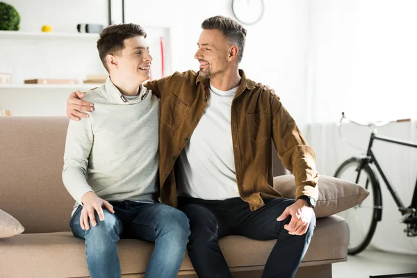 Glücklicher Vater und jugendlicher Sohn, die sich zu Hause auf dem Sofa umarmen und einander anschauen — Stockfoto