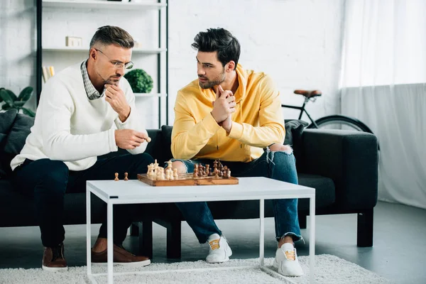 Nachdenklicher Sohn und reifer Vater spielen am Wochenende zu Hause gemeinsam Schach — Stockfoto