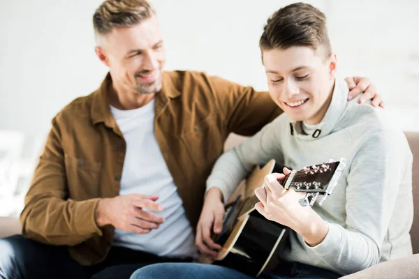 Selective focus of happy teen son playing acoustic guitar for father at home — Stock Photo