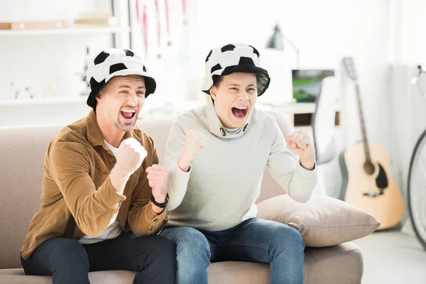 Excited father and teen son in football hats watching game and screaming on sofa at home — Stock Photo