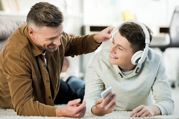 Sonriente hijo adolescente acostado en el suelo y mostrando algo en el teléfono inteligente al padre - foto de stock